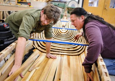 Tim Frandy (at left wearing green shirt), an outreach specialist at the UW Collaborative Center for Health Equity, helps, Wayne Valliere, (at right with ponytail) a Native American artist-in-residence with the Department of Art and member of the Lac du Flambeau Band of Lake Superior Chippewa Indians, install cedar wood slats and steam-bent ribs as Valliere constructs a traditional Ojibwe birchbark canoe at the Mosse Humanities Building at the University of Wisconsin-Madison.