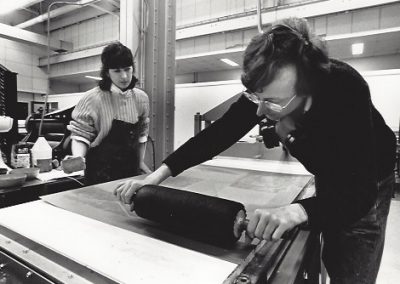 A professor demonstrates rolling out ink on a large plate for printing while a student watches.