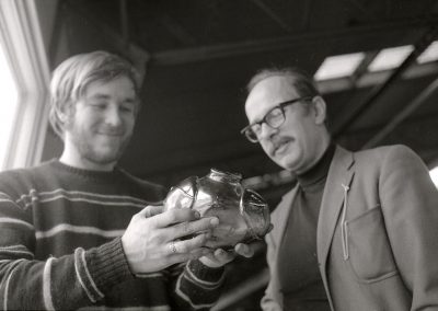 Professor of glass Harvey Littleton looks on while a student holds up a glass vase. Littleton taught at the University of Wisconsin-Madison from 1951 to 1976.
