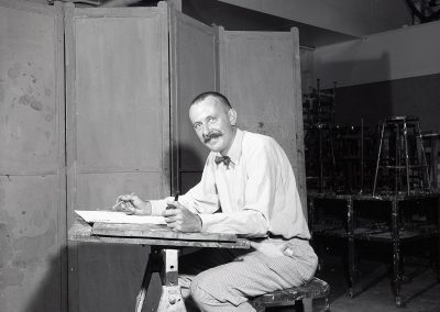 Professor John Wilde poses at a drafting table in September of 1960. Wilde was associated with the Magic Realism movement and Surrealism in the United States. His darkly humorous figurative imagery often included self-portraits through which he interacted with the people, animals and surreal objects that populated his fantasy world.