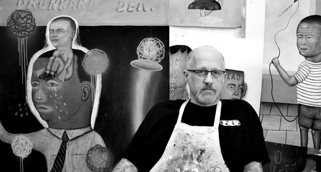 A black-and-white photo depicts Fred Stonehouse, lower in the frame and slightly right of center, looking at the camera in a black shirt and splattered art smock. Behind him are several of his visual art pieces. Each of these pieces are cartoonish in style and feature young men with slightly oversized heads. One is in tears and one has “A dingus” scrawled across his forehead, another flies a kite.