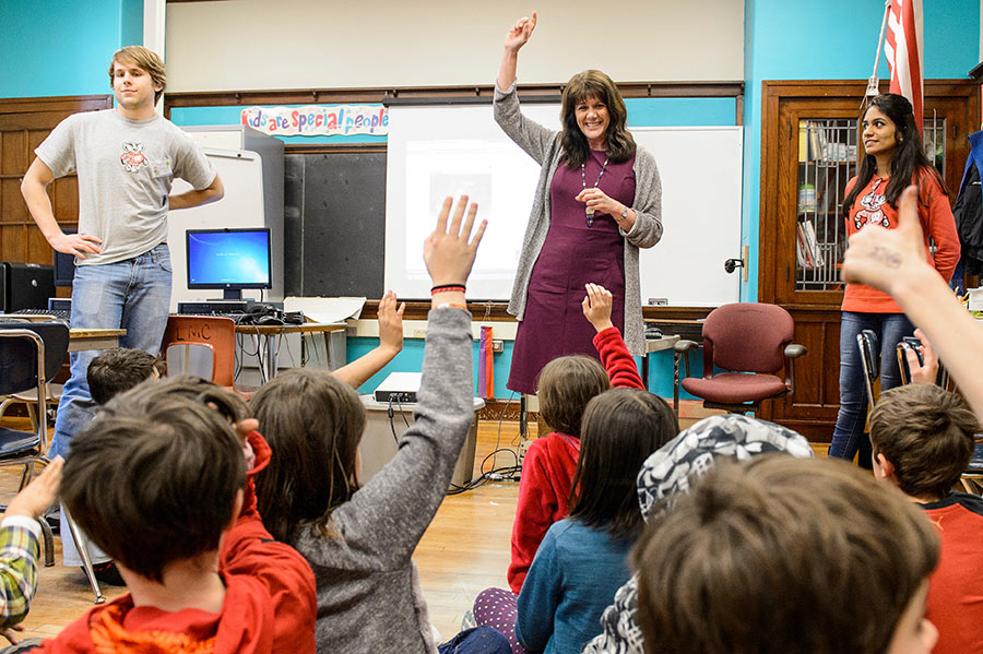 An instructional coach stands in front of a group of elementary school students who are seated on the floor and raising their hands.